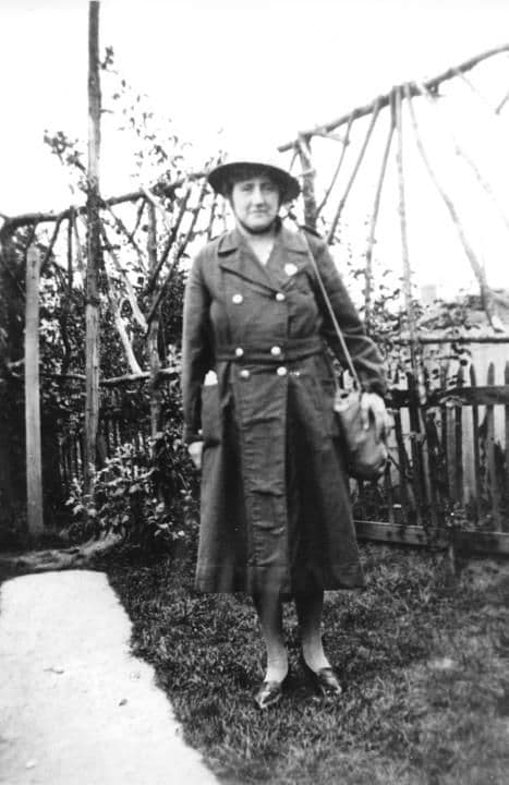 A woman in a Second World War air-raid warden's uniform, helmet and coat, standing in a garden.