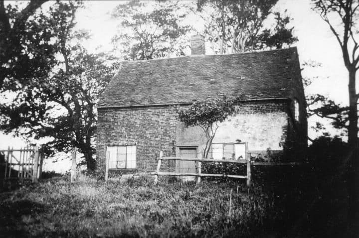 A small old brick cottage with a rustic fence in front, framed by trees.