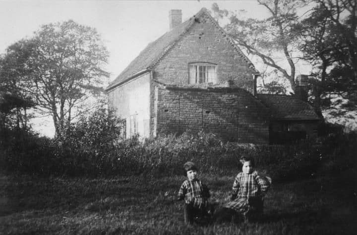 An old brick cottage among trees, with two small children sitting on the grass in front.