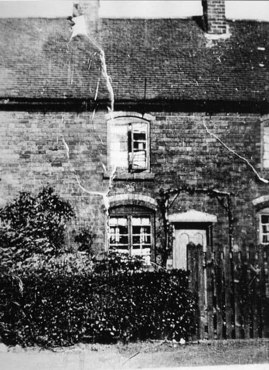 An old brick cottage with small windows and a creeper on its wall, behind a hedge and gate.