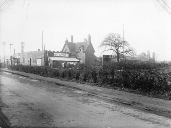 A muddy road with a hedge in the foreground and, behind it, a house and a single-storey shop with advertising signs.