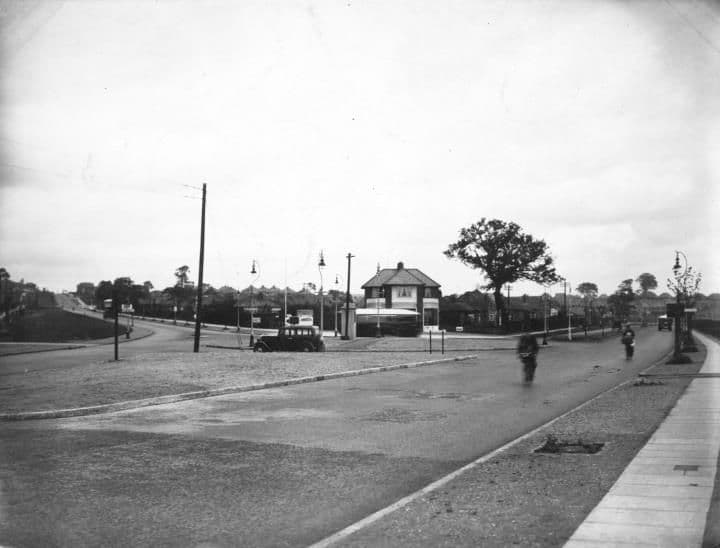 A wide road junction with a cyclist, a parked car and a small building in the distance, beside a broad grass verge.
