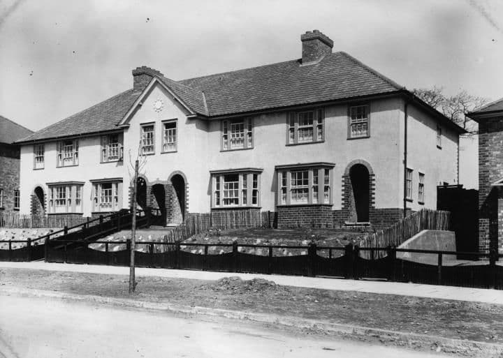 A newly built pair of rendered semi-detached houses with a central gable, round-arched brick porches and bay windows, behind new timber fencing.
