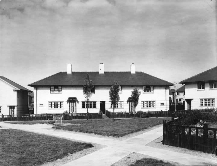A terrace of white rendered houses with three chimneys behind a lawn crossed by paths, with young trees.