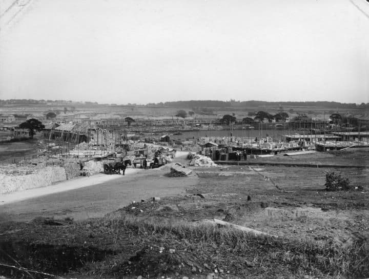 An open building site with houses under construction, rough ground in the foreground and trees on the distant skyline.