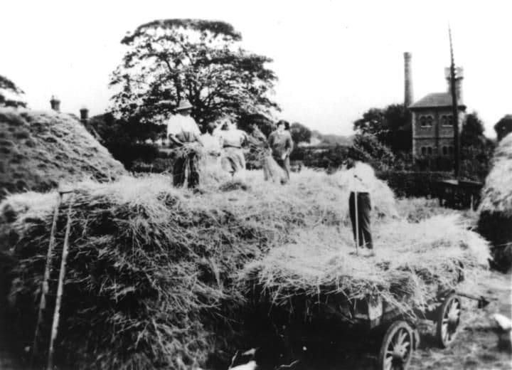 Farm workers loading hay onto a cart, with a towered brick building and a tall chimney behind.