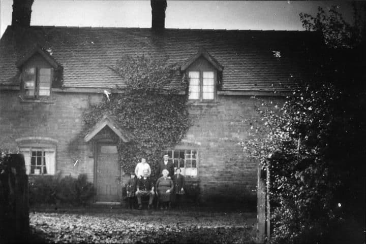 A brick farmhouse with dormer windows and a creeper-covered porch, with a small group of people seated in front.