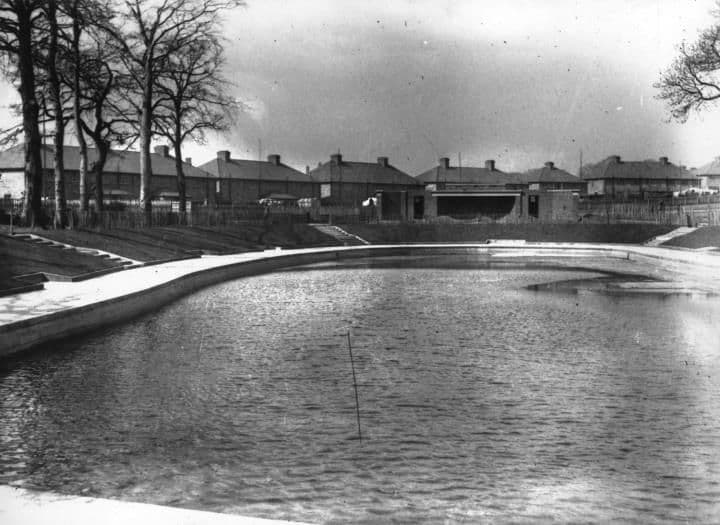 A large pool of water with a curved concrete edge, with rows of houses and bare trees behind.