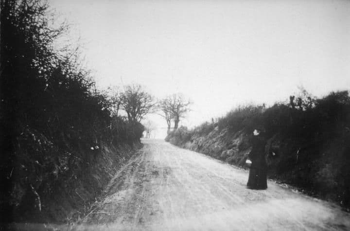 A sunken country lane between high hedges, with a woman in a long dark dress standing at the side.