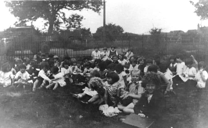 A large group of children sitting together on grass beneath trees.
