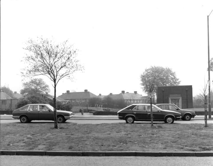A small flat-roofed brick building beyond a grass verge with young trees, with 1980s cars parked on the road and houses behind.