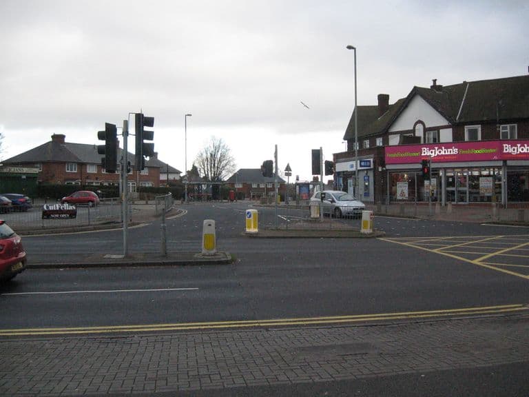 A road junction with traffic lights looking over to Warren Farm Road, with a parade of shops on the right.