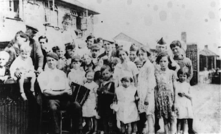 A large crowd of children and adults posed in a street in front of houses, one man holding an accordion.