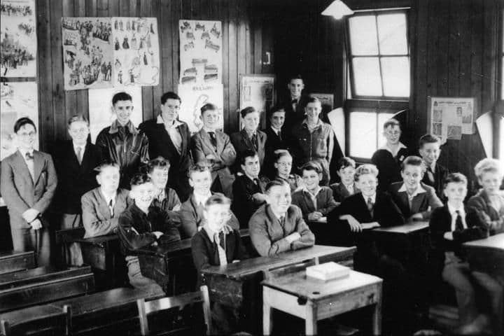 Teenage boys seated at wooden desks in a classroom with posters on the wall.