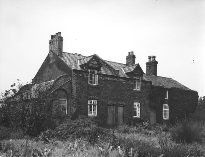 A two-storey brick house with dormer gables, tall chimneys and a creeper-covered end, in an overgrown garden.