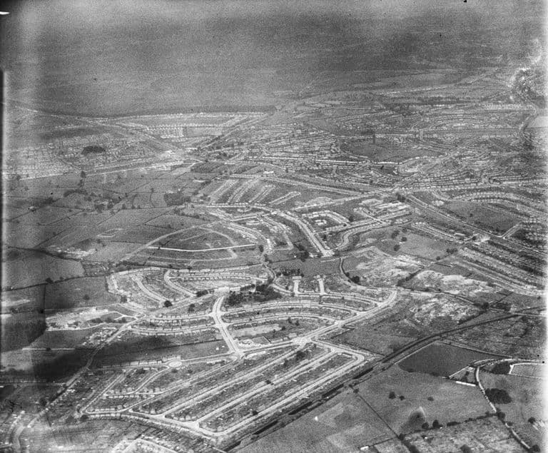 Oblique aerial photograph from the south-west: newly laid-out curving streets of houses in the foreground, some unfinished, with the older estate beyond.