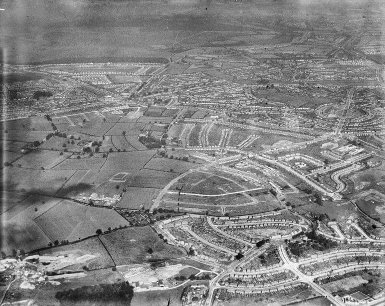 Oblique aerial photograph from the south-west: curving terraces of new houses among fields, with the built-up estate stretching to the horizon.