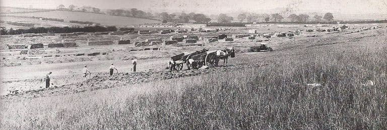 A wide panorama of open field being prepared for building: workmen with hand tools, horse-drawn carts and a lorry, with materials stacked in rows and hedgerow trees on the skyline.