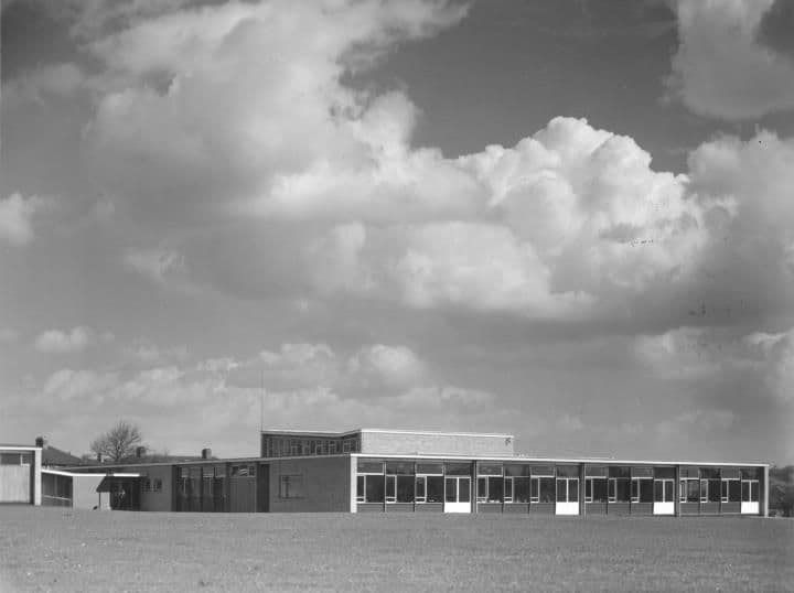 A long flat-roofed modern school building on open grass under a cloudy sky.