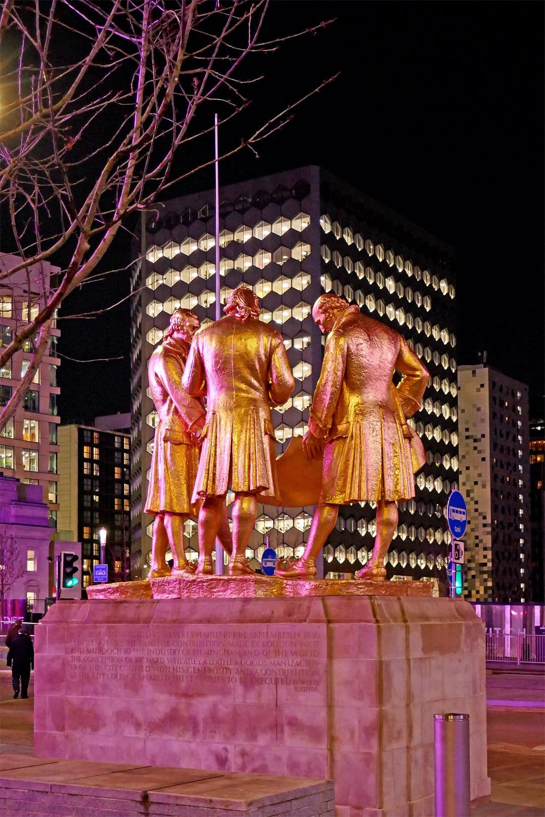 Gilded bronze statue of three men in eighteenth-century dress studying a drawing, on a stone plinth in a city square.