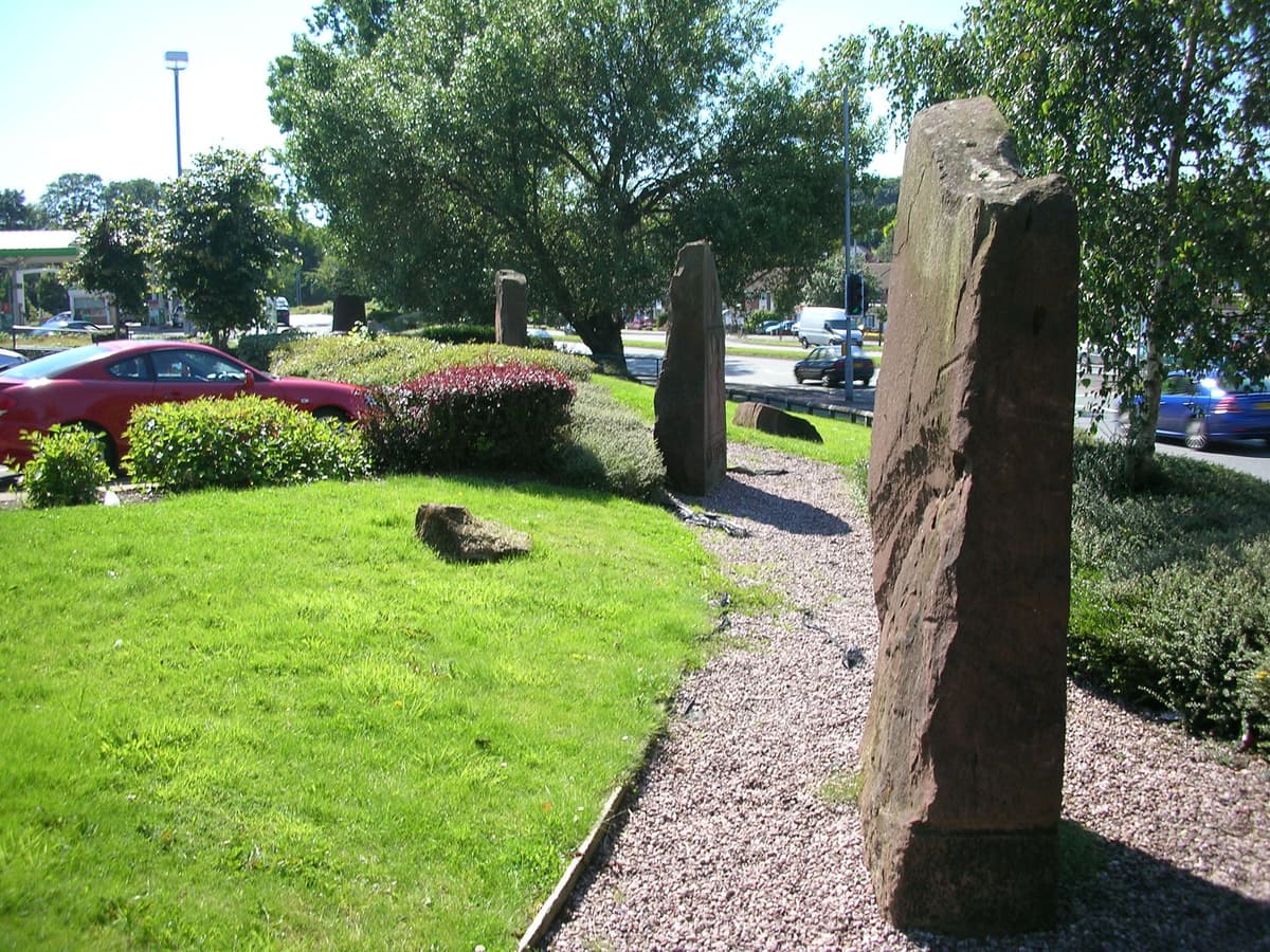 Carved sandstone memorial stones standing in a row on grass beside a car park.