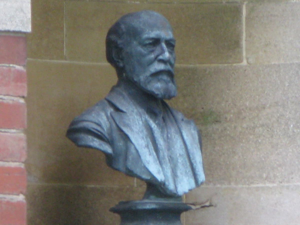 Stone bust of an elderly bearded man in an arched alcove in a brick wall.