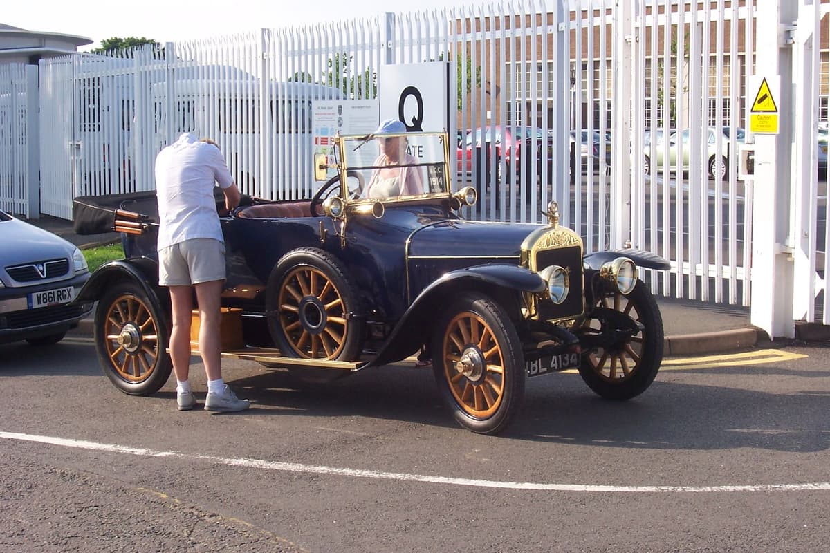 A dark blue veteran open tourer with brass lamps parked by a works gate marked with a large letter Q.