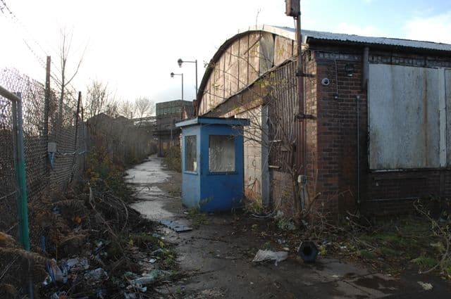 A rubbish-strewn path beside a fence, a blue security hut and an old brick and corrugated-metal works building.