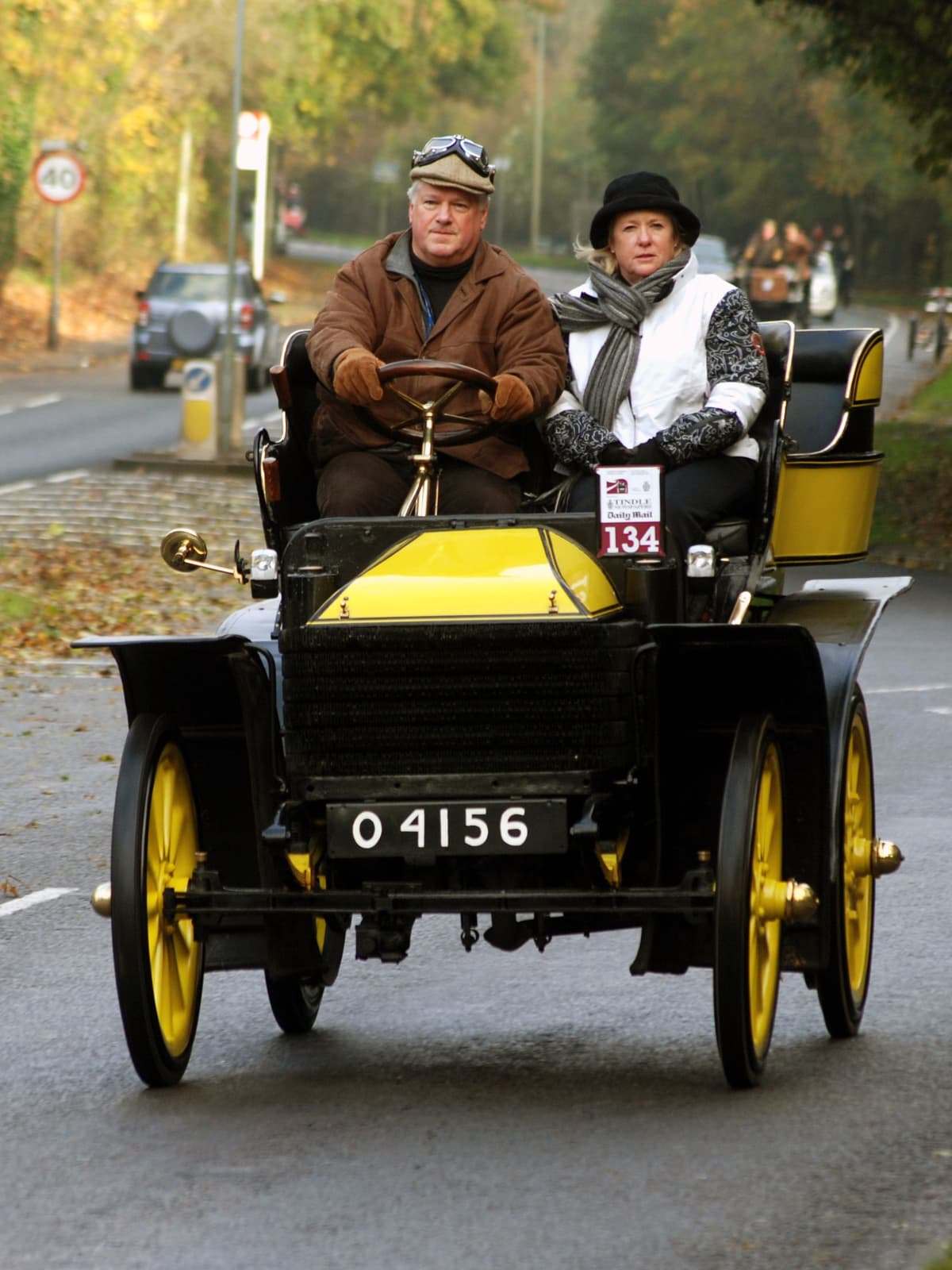 A black and yellow veteran car carrying a man and a woman along a road during the London to Brighton run.