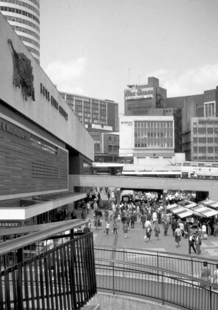1960s photograph of a large concrete shopping centre beside market stalls under a road.
