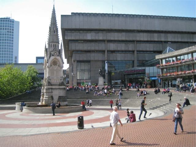Gothic memorial fountain in a square, with a large concrete library behind.