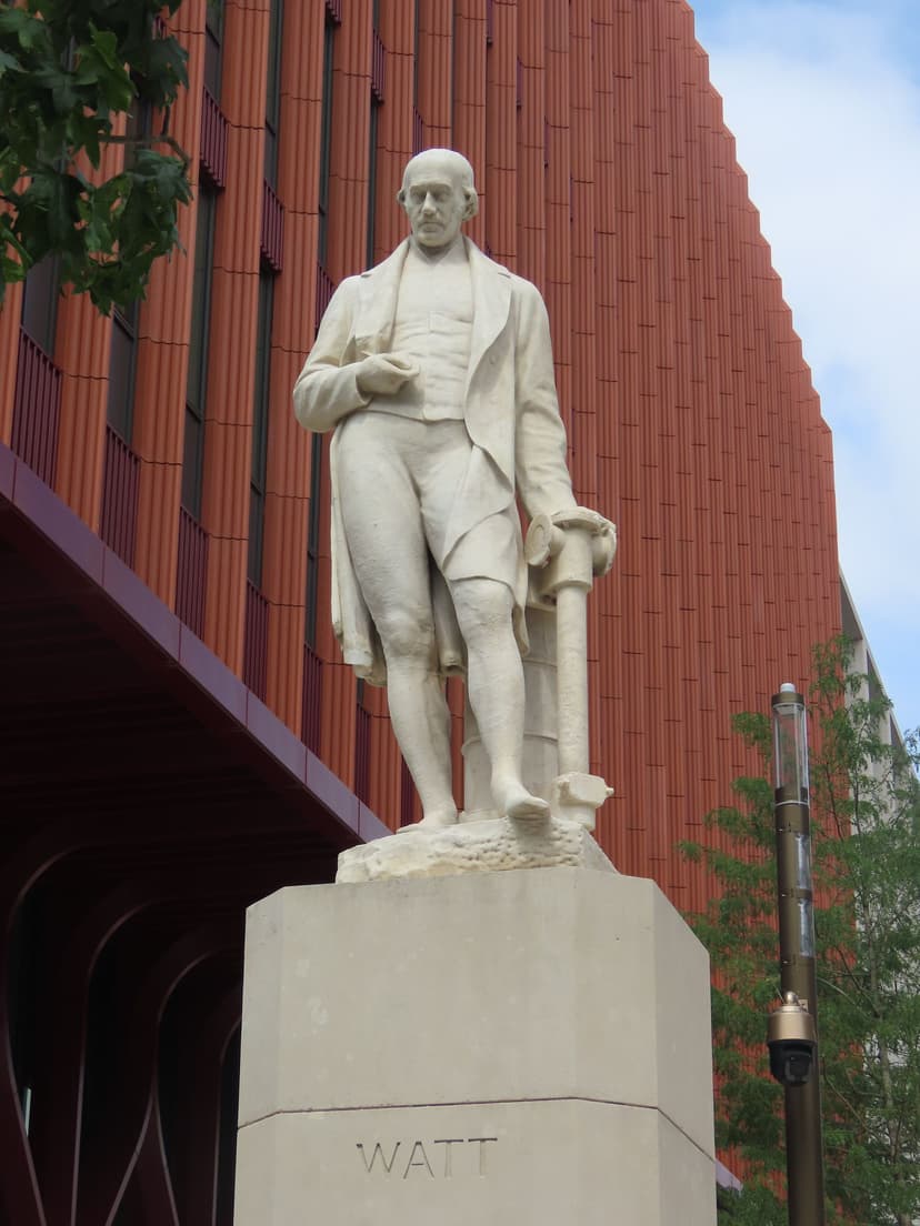 A pale stone statue of a man in a long coat holding compasses, on a tall plinth beside a new walkway.