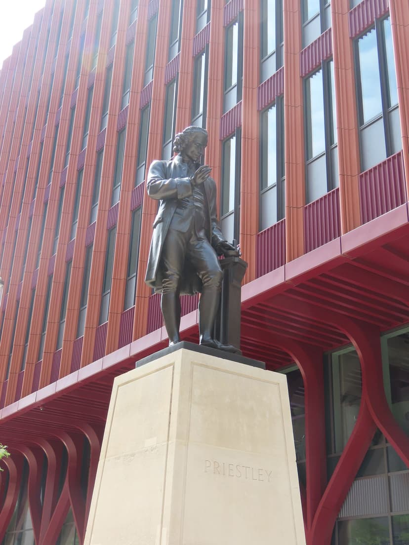 A dark bronze statue of a man in eighteenth-century dress on a tall stone plinth beside a walkway.