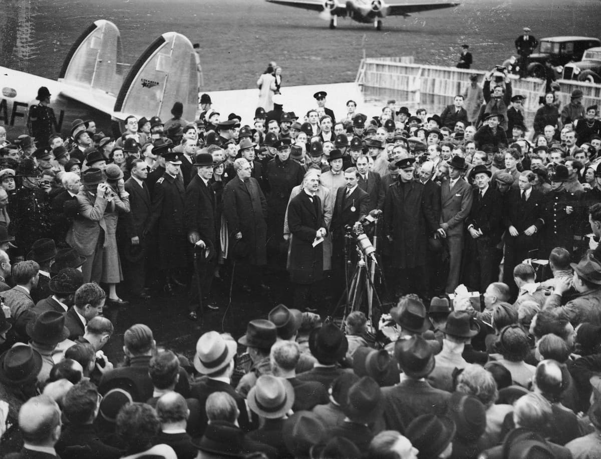 A dense crowd in hats and coats surrounds a group of men and a newsreel camera at an aerodrome, with an aircraft behind.