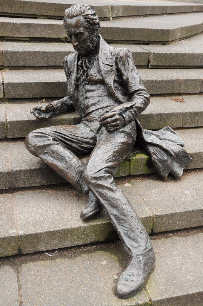 A bronze figure of a man in a long coat sitting on stone steps, a sheet of paper in his hand.