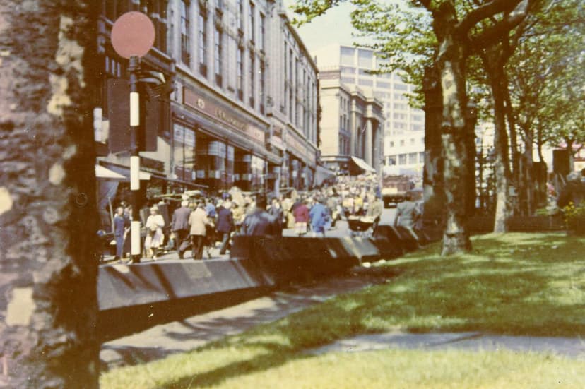 Colour photograph of shoppers crowding a street of tall Victorian shops beside a grassy churchyard, looking up a slope.