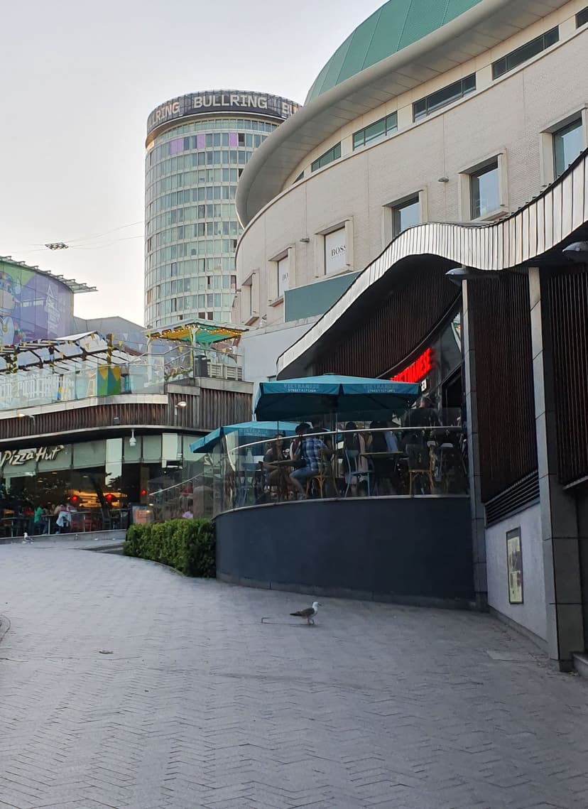 A paved street sloping past restaurant terraces towards the cylindrical Rotunda tower, with the Bullring centre’s curved wall on the right.