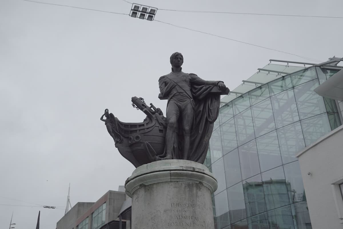 Bronze statue of Nelson beside a ship’s prow on a plain round stone pedestal, with a modern glass building behind.