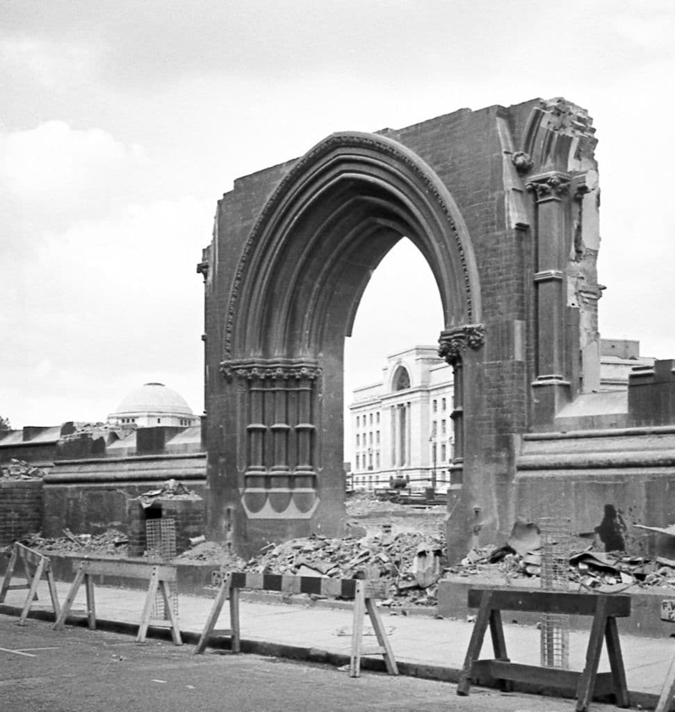 A ruined Gothic archway on a demolition site, with a white civic building and a dome visible beyond.