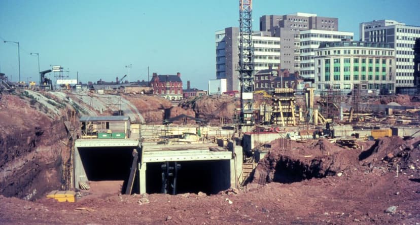 Colour photograph of a concrete road tunnel portal under construction amid raw earthworks, with office blocks behind.