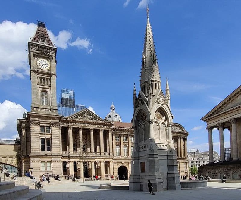 Sunny paved square with the Gothic Chamberlain Memorial, the clock tower of the Council House and the museum, and the columns of the Town Hall on the right.