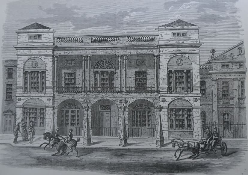 Engraving of a classical theatre front with arched doorways, paired columns and a balustrade, horses and a carriage in the street.