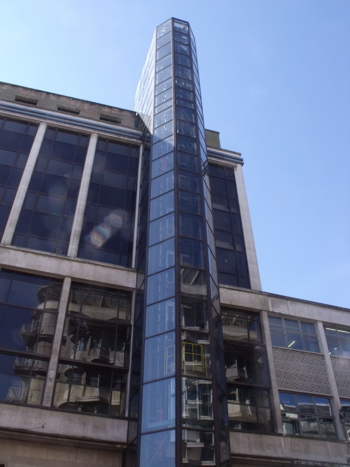 A tall glass lift tower attached to a post-war commercial building on a shopping street.