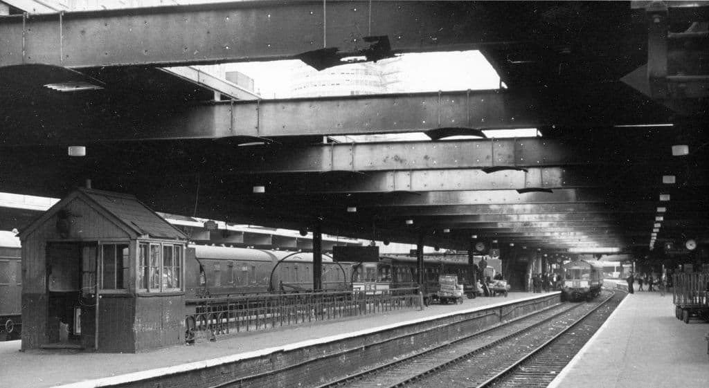 Photograph of station platforms under old roof girders, with a round office tower glimpsed beyond.