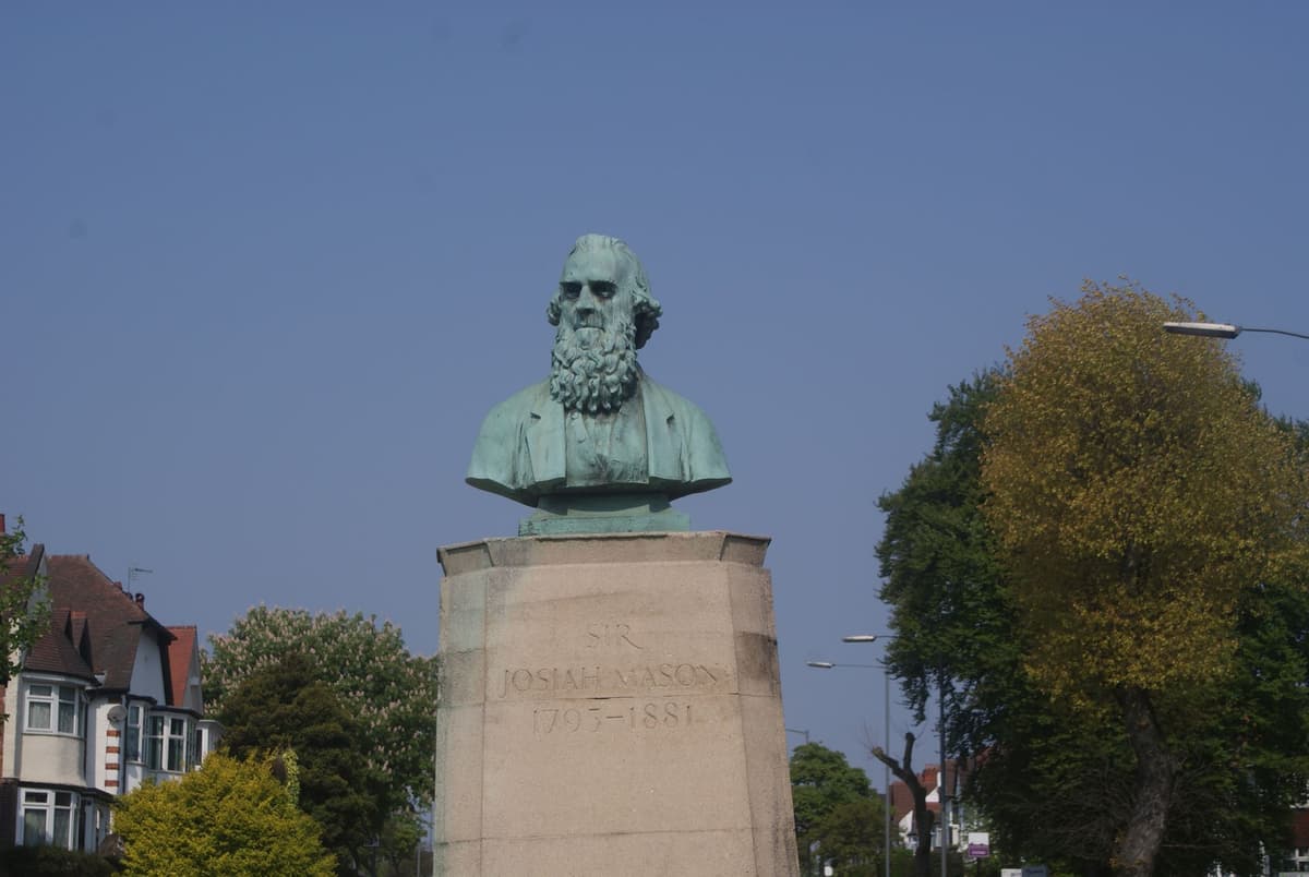 Green bronze bust of a bearded old man on a stone pedestal inscribed Josiah Mason 1795–1881, with trees and houses behind.