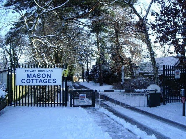 Snow-covered gated entrance with a sign reading Private Grounds, Mason Cottages, and bungalows among trees beyond.
