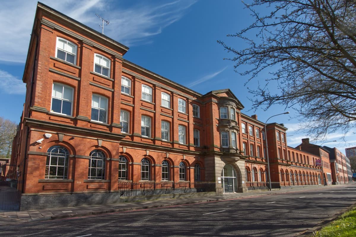 Long three-storey red-brick building with round-arched ground-floor windows, a stone arched entrance and a bow window above it, seen along a road under blue sky.