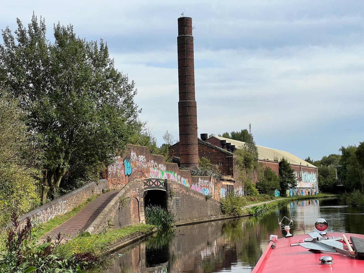 A tall brick chimney and old brick industrial buildings beside a canal, with a towpath bridge in front.