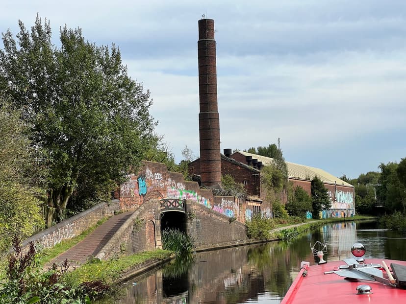 A tall brick chimney and old brick industrial buildings beside a canal, with a towpath bridge in front.