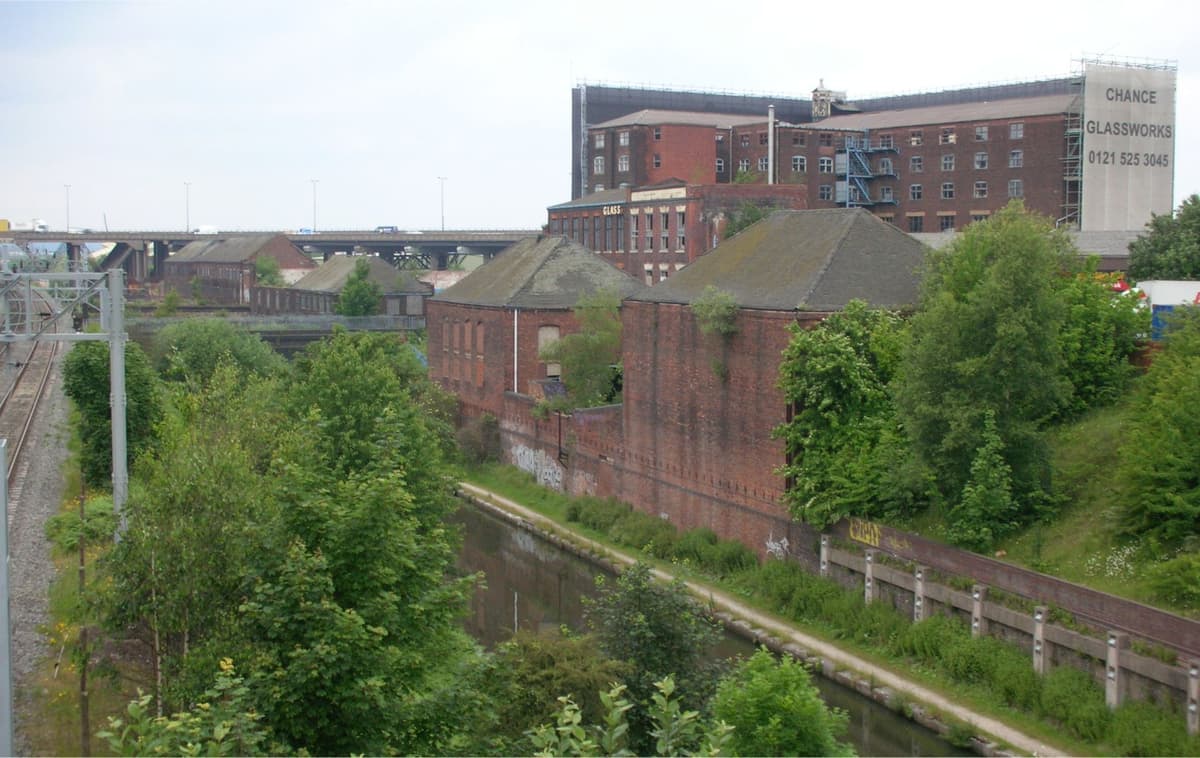 Long range of old brick industrial buildings beside a canal, with a railway alongside and a motorway viaduct behind.